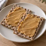 Två bitar knäckebröd med ett tunt lager ljusbrunt messmör på en benvit tallrik.